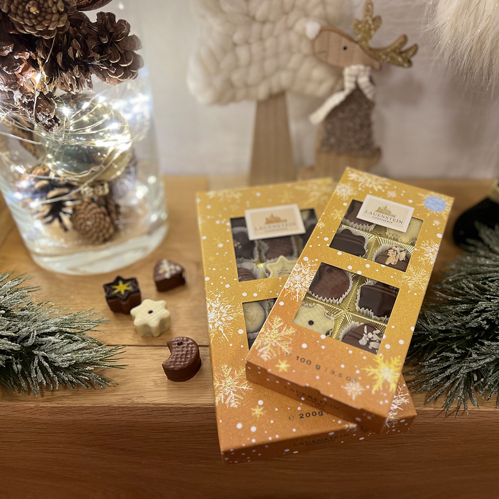 Zwei Weihnachts-Auslesen (100g und 200g) auf Holz, umrahmt von weihnachtlicher Deko und Pralinen