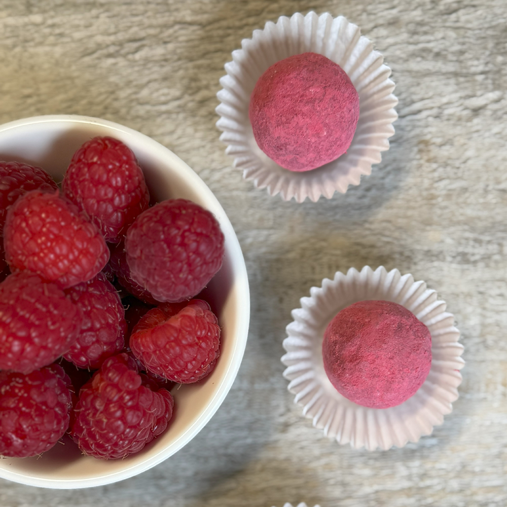 Rosa Himbeertrüffel in weißen Papierförmchen neben einer Schale mit frischen Himbeeren