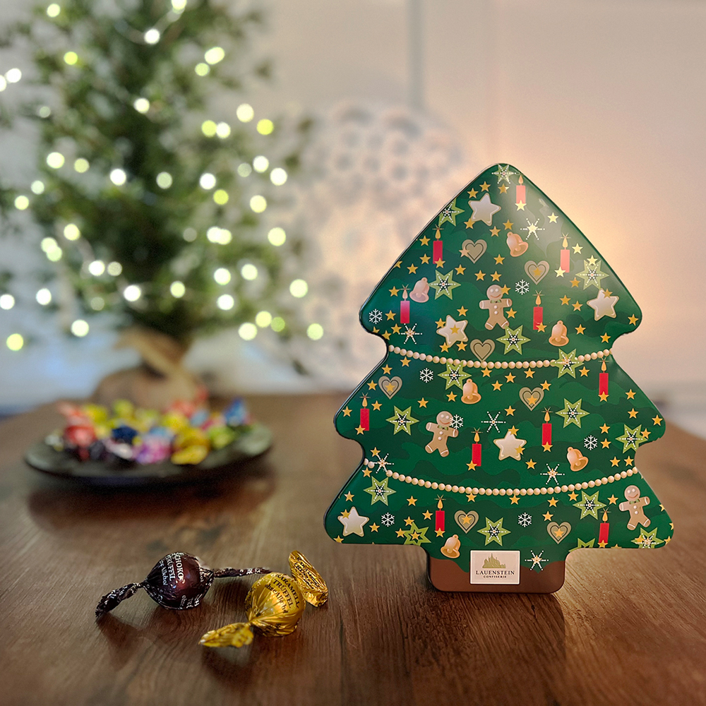 Aufrecht stehende Metalldose auf Holztisch mit bunt gewickelten Gourmetkugeln vor einem warm leuchtenden Weihnachtsbaum