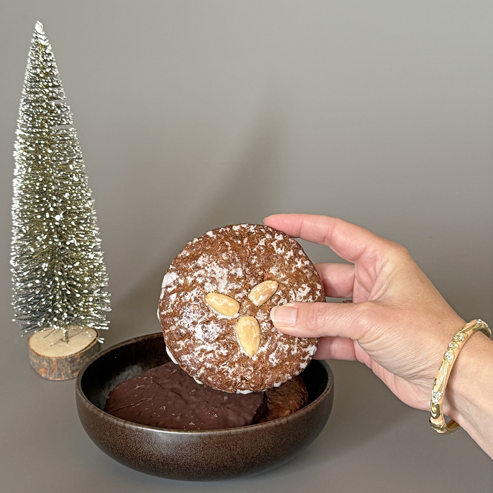 eine Hand hält einen Elisen-Lebkuchen mit Mandeln über einer Schale mit Schoko-Lebkuchen und Weihnachtsdeko