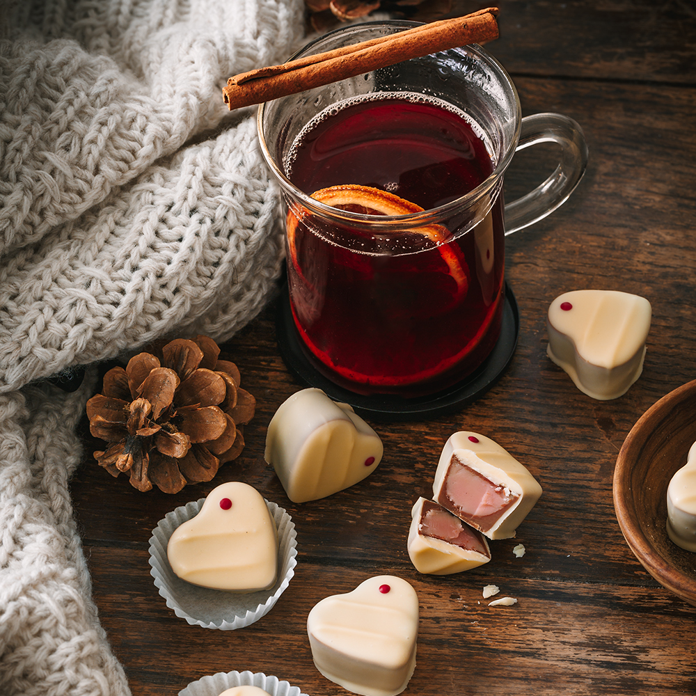 weiße Pralinen in Herzform mit heller Füllung neben einem Glas Glühwein, einer Zimtstange und Deko