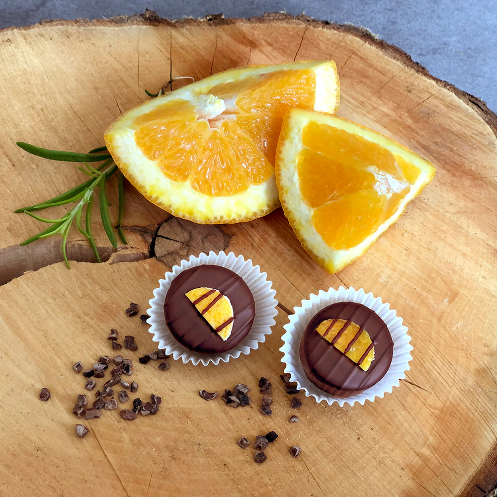 Zwei dunkle Schokopralinen mit kandierten Orangenspalten und Schokostreifen auf einer Baumscheibe garniert mit Kakaonibs