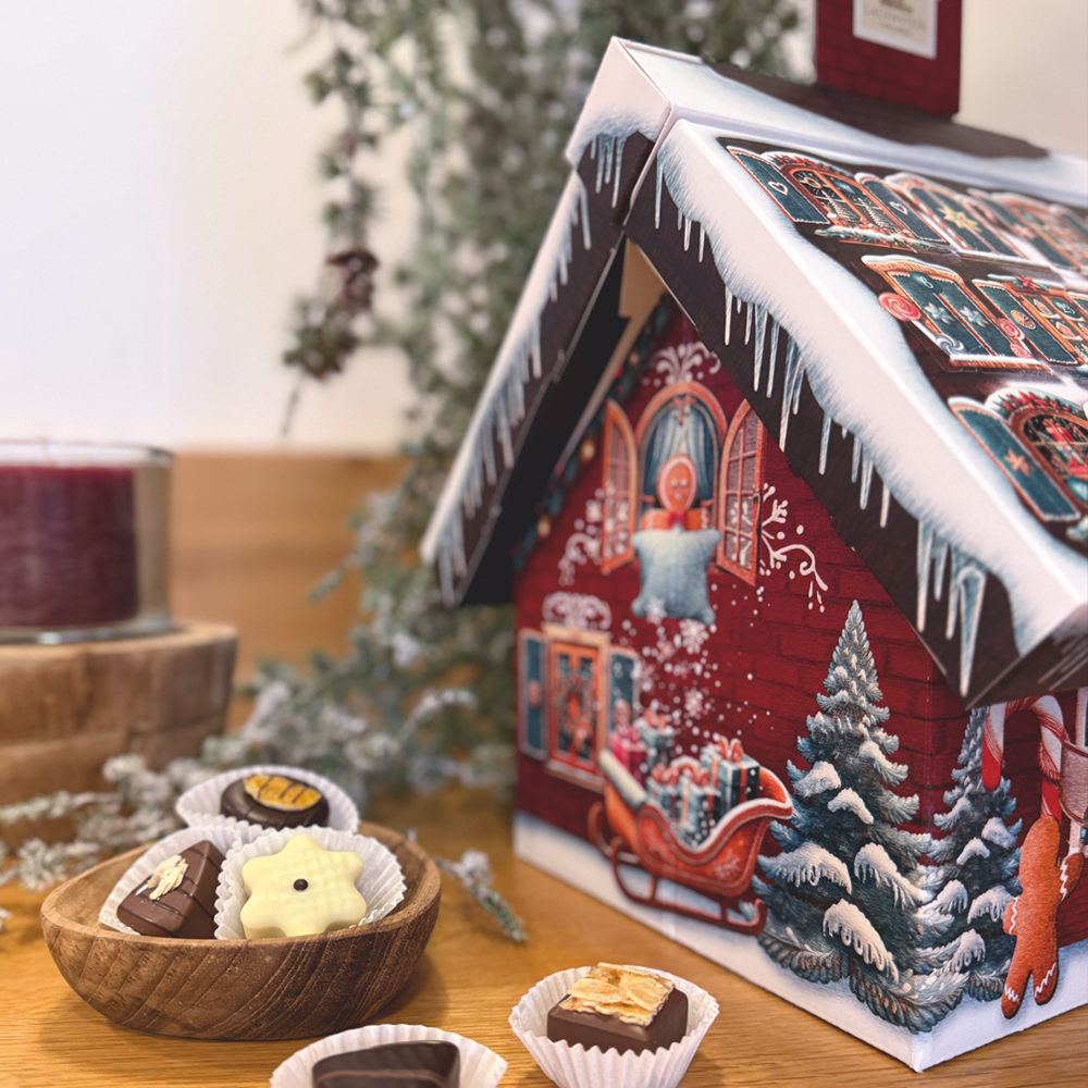 Adventskalenderhaus neben einer Holzschale mit Pralinen vor weihnachtlicher Deko