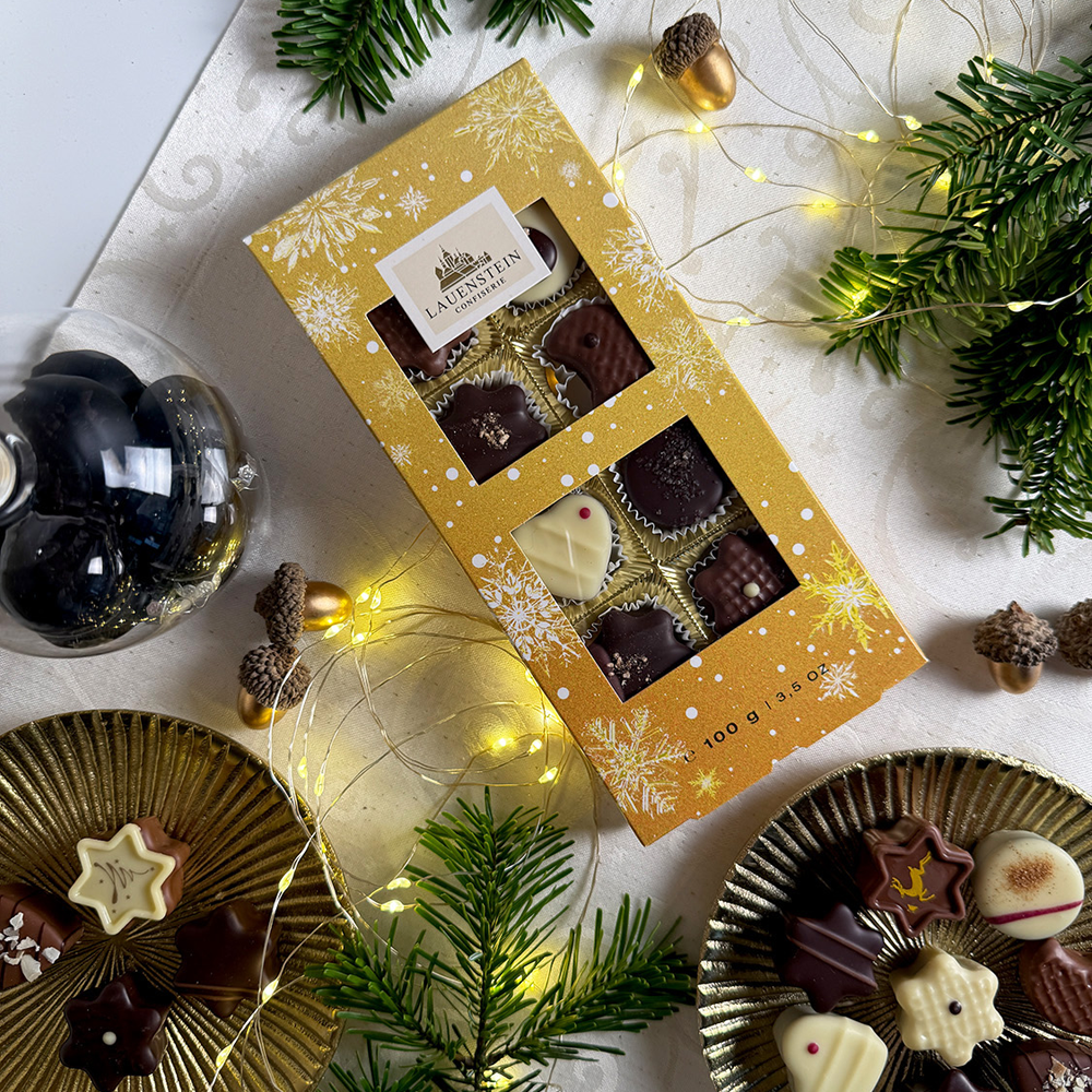 Pralinenschachtel umgeben von Tannengrün, einer Lichterkette und feinen Pralinen auf Goldtellern