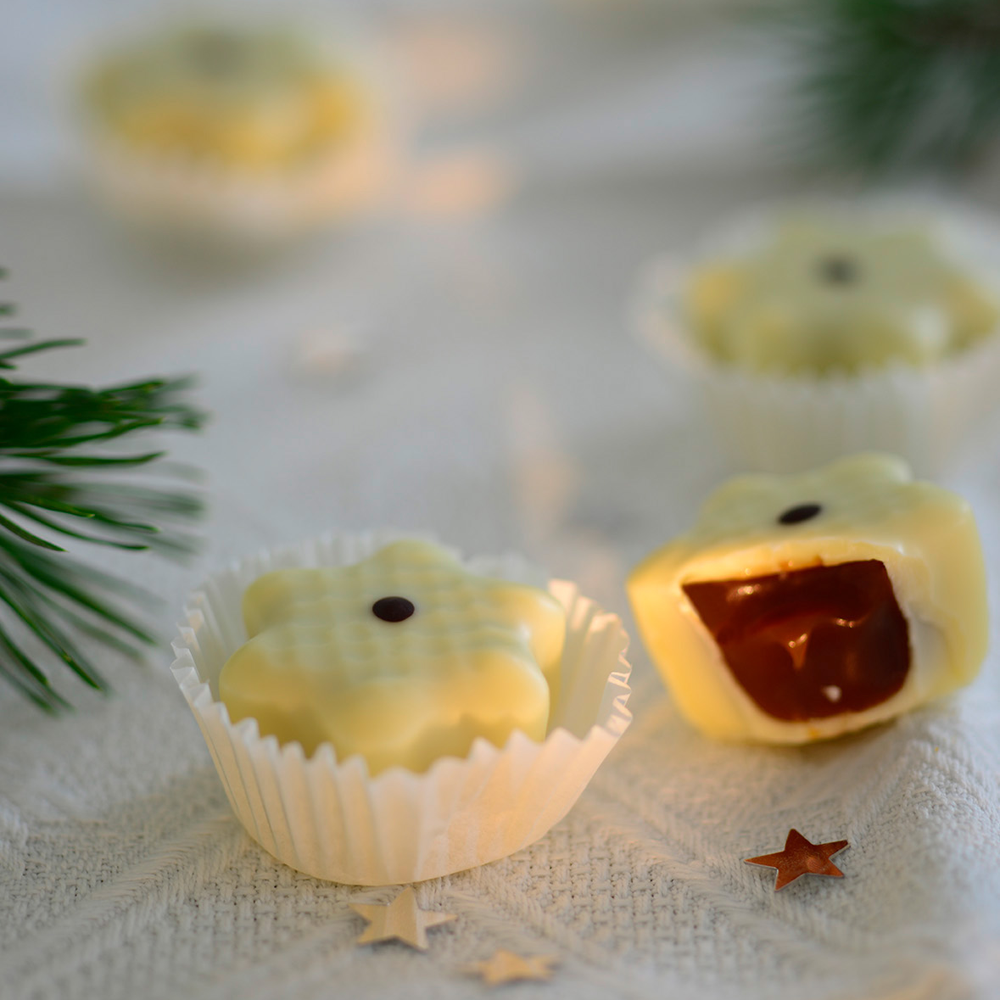vier weiße Stern-Pralinen, eine davon angeschnitten mit dunkler Cremefüllung auf weißem Deckchen mit weihnachtlicher Dekoration