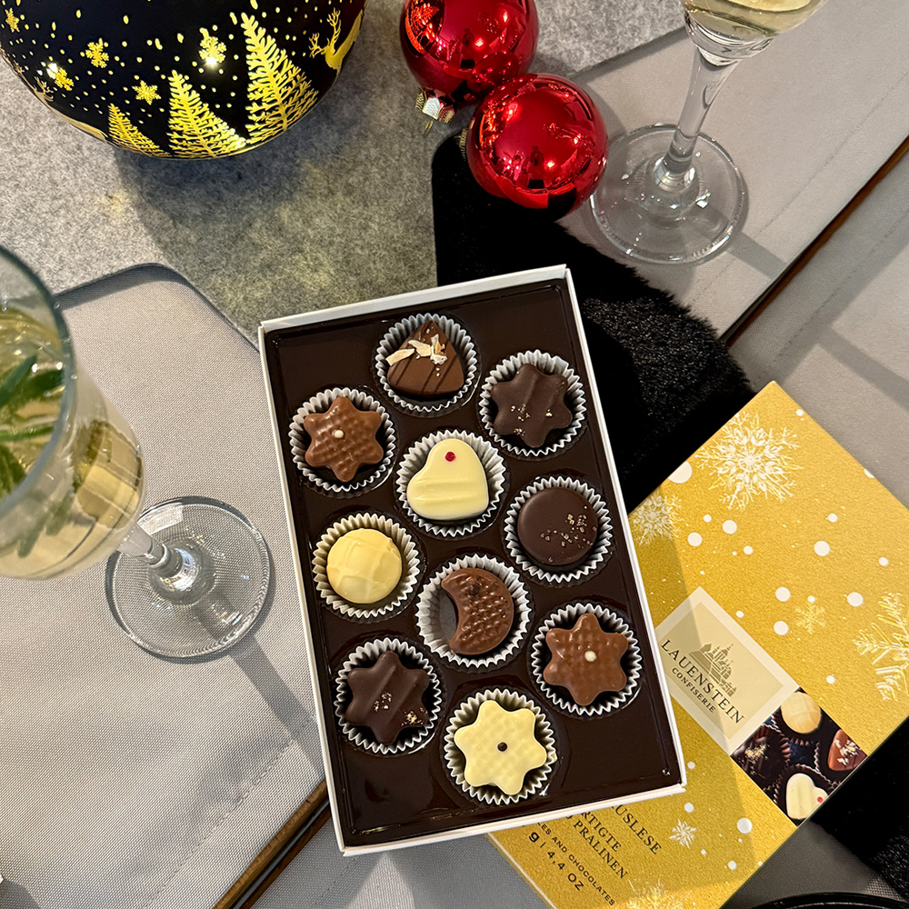 Festlich gedeckter Tisch mit geöffneter Pralinen-Auslese, zwei Sektgläsern und rotem Weihnachtsschmuck